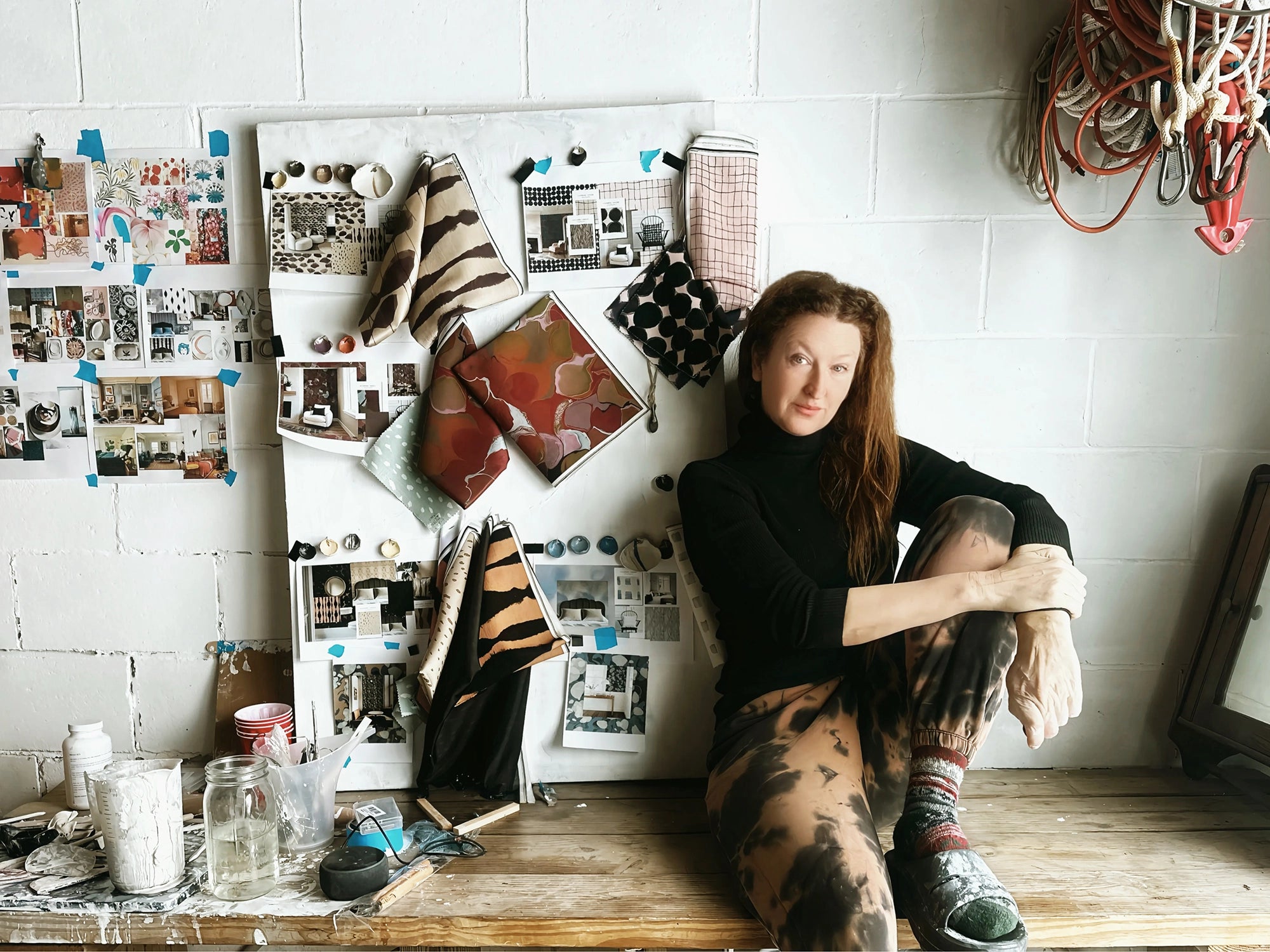 female artist sitting on a wooden table with a white wall and home decor artwork and pottery studio items in the background